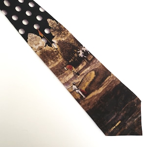 May include: A black necktie with a golf-themed design. The top portion features white polka dots, while the lower section depicts a golf course scene with golfers in action. The tie is made of a smooth fabric.