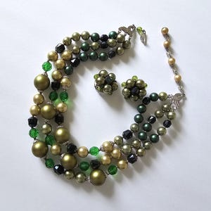 May include: A vintage three-strand necklace and matching clip-on earrings set. The necklace features olive green, black, and gold-toned beads, with green and black crystal accents. The earrings have a cluster design with similar colours. The set is on a white background.