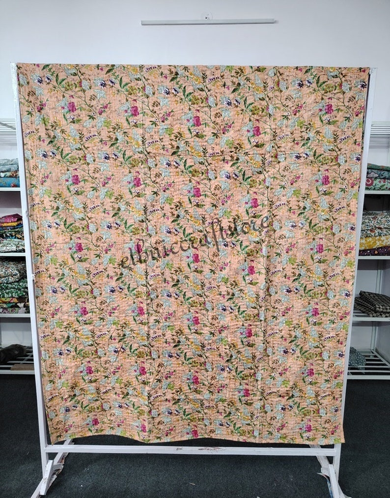 May include: A peach-colored kantha quilt with a floral pattern. The quilt is made of cotton and is hand-stitched. It is a traditional Indian textile.