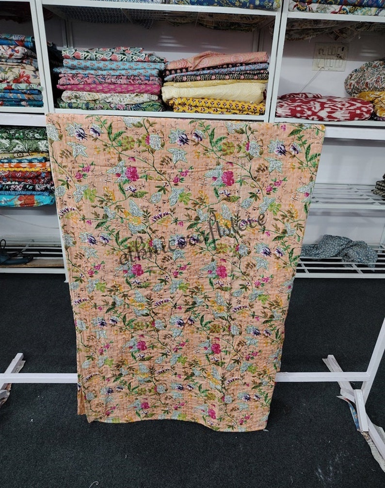 May include: A peach-colored kantha quilt with a floral pattern. The quilt is made of cotton and is hand-stitched. It is a traditional Indian textile.