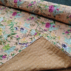 May include: A peach-colored kantha quilt with a floral pattern. The quilt is made of cotton and has a hand-stitched design. The quilt is folded in half and the stitching is visible on the back.