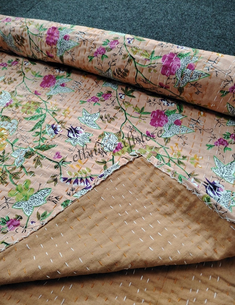 May include: A peach-colored kantha quilt with a floral pattern. The quilt is made of cotton and has a hand-stitched design. The quilt is folded in half and the stitching is visible on the back.