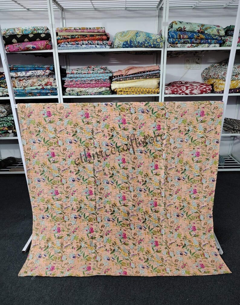 May include: A peach-colored kantha quilt with a floral pattern. The quilt is made of cotton and is hand-stitched. It is a traditional Indian textile.
