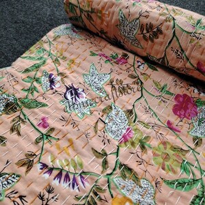 May include: A peach-colored kantha quilt with a floral pattern in shades of green, white, pink, and yellow. The quilt is hand-stitched with a grid pattern.