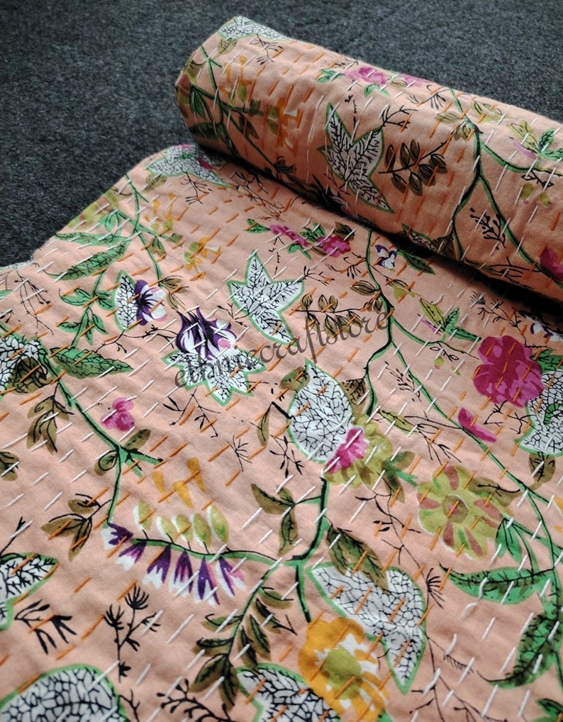May include: A peach-colored kantha quilt with a floral pattern in shades of green, white, pink, and yellow. The quilt is hand-stitched with a grid pattern.