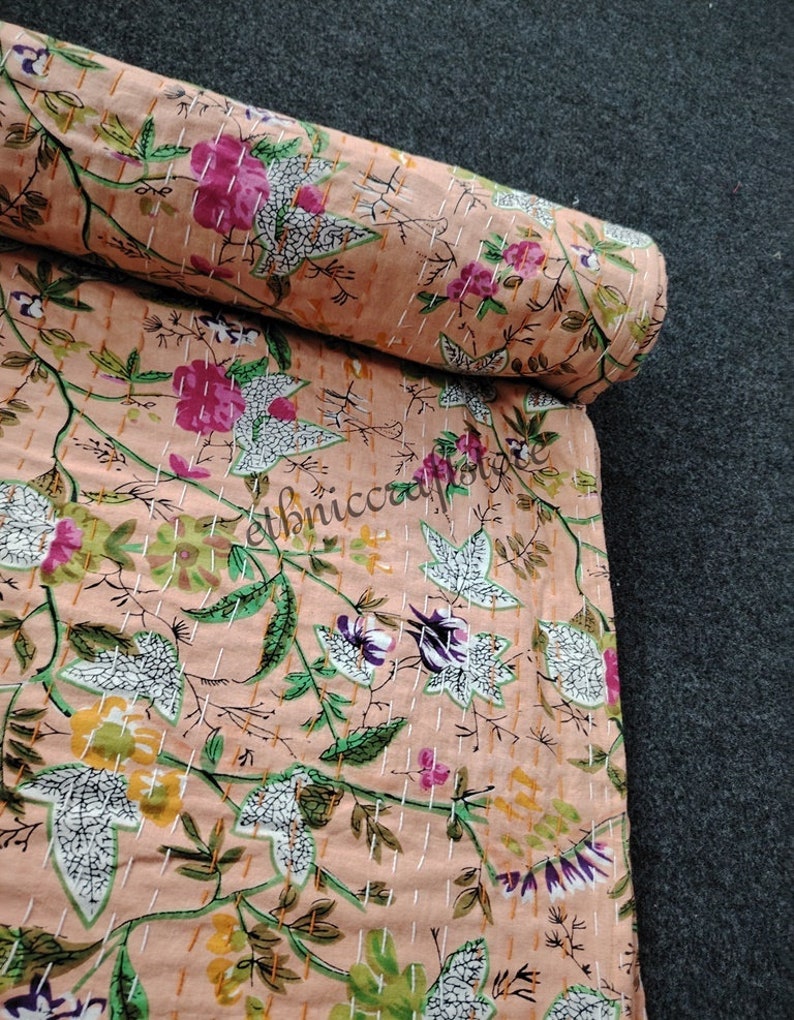 May include: A peach-colored fabric with a floral print featuring white, pink, green, and yellow flowers. The fabric is rolled up and has a white stitching pattern.