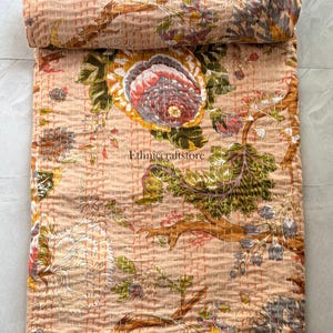 May include: A peach-colored cotton quilt featuring a floral pattern with large blossoms in shades of pink, yellow, and green, accented by brown branches. The fabric is detailed with visible orange running stitches and the text Ethniccraftstore.