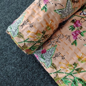 May include: A close-up of a peach-colored fabric with a floral pattern. The fabric is quilted with white stitching and has a subtle texture. The floral pattern features pink flowers, green leaves, and white accents.