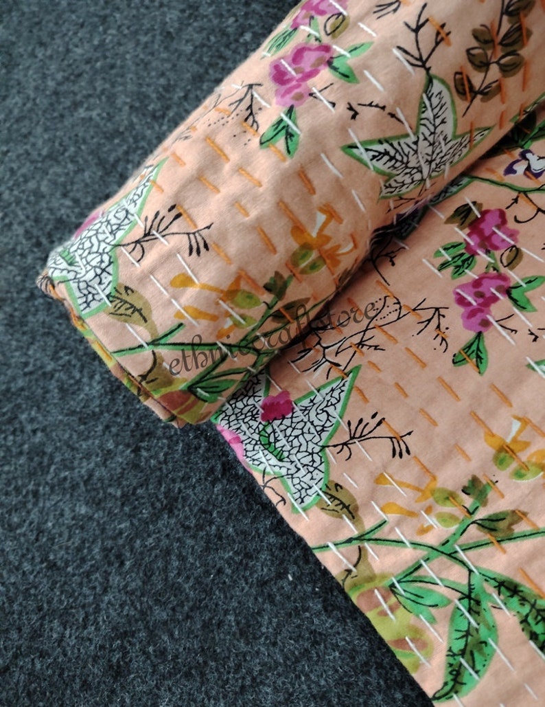 May include: A close-up of a peach-colored fabric with a floral pattern. The fabric is quilted with white stitching and has a subtle texture. The floral pattern features pink flowers, green leaves, and white accents.