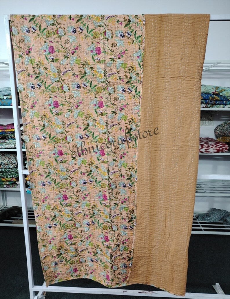 May include: A peach-colored kantha quilt with a floral print and a solid brown back. The quilt is hand-stitched and has a vintage look.