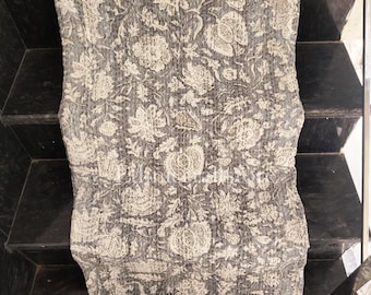 May include: A gray and white floral patterned quilt. The quilt features a repeating design of stylized flowers and foliage. The stitching is visible, adding texture to the fabric. The quilt is folded, showcasing its design and construction.