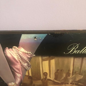 May include: A framed artwork featuring a ballet theme. The image showcases a ballerina's foot in a pink satin pointe shoe adorned with pearls and gold bracelets. The background depicts a ballet studio with dancers.