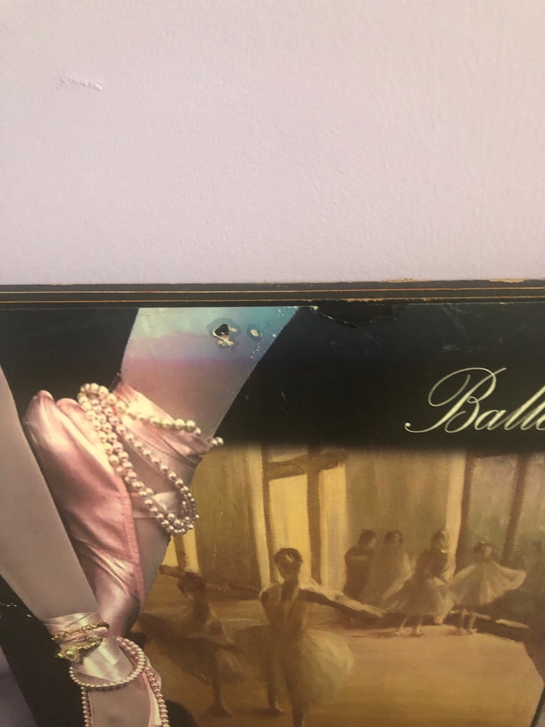 May include: A framed artwork featuring a ballet theme. The image showcases a ballerina's foot in a pink satin pointe shoe adorned with pearls and gold bracelets. The background depicts a ballet studio with dancers.
