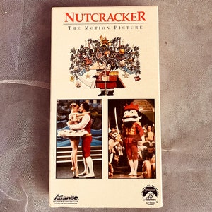 May include: A vintage VHS tape for "Nutcracker: The Motion Picture." The cover features illustrations of the Nutcracker, a ballet scene, and a stage performance. The title is in red, and the tape is from Atlantic Releasing Corporation.