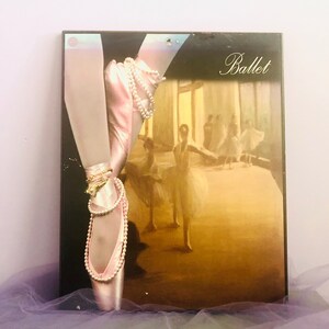 May include: A framed artwork featuring a ballet dancer's pointe shoe with a pearl necklace and bracelet. The artwork has the word "Ballet" in script. The background shows a ballet performance with dancers in white tutus.
