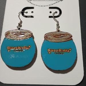 Könnte beinhalten: Handgefertigte Ohrringe mit türkisblauem Design und dem Schriftzug "BuzzBallz Cocktails" in Gold. Die Ohrringe sind wie die Getränkebehälter geformt und haben silberfarbene Ohrhaken. Auf einer weißen Karte mit dem Text "J's Jewelry" präsentiert.