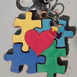 May include: A wooden puzzle keychain with four puzzle pieces, a red heart, and a small puzzle piece charm. The puzzle pieces are painted blue, green, yellow, and red. The keychain is attached to a silver metal ring.