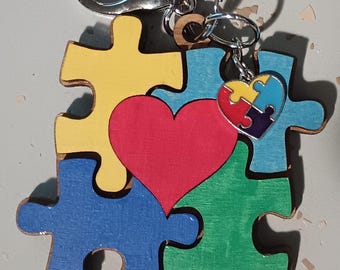 May include: A wooden keychain featuring a puzzle piece design with a red heart in the center. The puzzle pieces are painted in yellow, blue, and green. A small heart-shaped charm with puzzle piece colors hangs from the keychain.