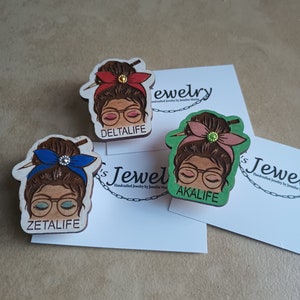 May include: Three wooden pins featuring a cartoon woman's face, each with a different coloured headband and a different name: "ZETALIFE", "DELTALIFE", and "AKALIFE".