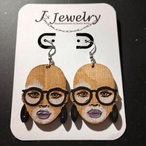 May include: A pair of wooden earrings featuring a hand-painted portrait of a person wearing glasses. The earrings are brown with black accents and have silver hooks.