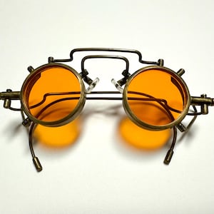 May include: Round glasses featuring bright orange lenses and a metallic bronze-toned frame. The design includes a prominent bridge, articulated side hinges, and curved temple arms with small cylindrical tips, creating a distinct mechanical appearance.