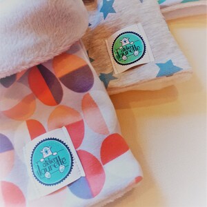 Peut inclure: Trois langes à  rot  pour bébé en tissu avec des motifs différents. Le premier est blanc avec une bordure en fourrure blanche, le second est gris avec des étoiles blanches et le troisième est blanc avec un motif géométrique coloré. Les trois langes ont une étiquette circulaire avec un ours polaire et le texte "atelier laurette".