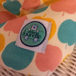 Peut inclure: Un gros plan sur un tissu jaune, rose et vert avec une étiquette blanche qui dit "atelier laurette" et un ours en dessin animé. Le tissu a un motif de pommes.