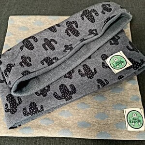 Peut inclure: Deux carrés de tissu avec des motifs différents. Le carré du haut est gris avec un imprimé de cactus noir. Le carré du bas est beige avec un imprimé de nuage bleu clair. Les deux carrés ont une petite étiquette blanche avec le texte "alben lourelle" en noir.