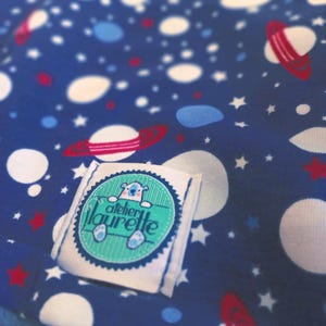 Peut inclure: Gros plan sur un tissu bleu avec un motif spatial comprenant des planètes blanches, des anneaux rouges, des étoiles blanches et des accents bleus. Une étiquette blanche avec le texte "atelier laurette" et un ours de dessin animé est cousue sur le tissu.