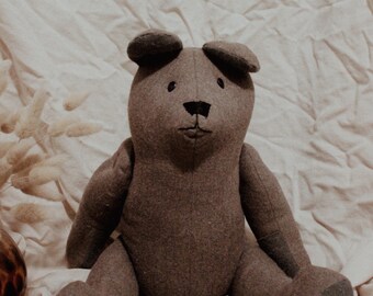 May include: A gray plush teddy bear with black eyes and a black nose sits on a white fabric background.