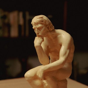 May include: A cream-colored sculpture of a seated figure, with a hand resting on the chin in a thoughtful pose. The figure is nude and sits atop a round base. The sculpture is lit from above, creating shadows.