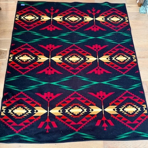 May include: A black blanket with a geometric pattern in red, gold, and green. The design features diamond and arrow shapes, creating a Southwestern or Native American aesthetic. The blanket is rectangular and appears to be made of a soft material.