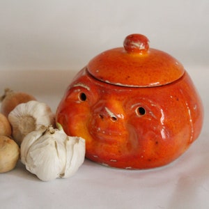 Op de afbeelding: Een oranje keramische knoflookpot met een deksel in de vorm van een gezicht. De pot heeft gaten voor luchtcirculatie. Er liggen vier teentjes knoflook en twee uien naast de pot.