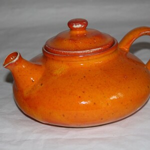 Puede incluir: Una pequeña tetera de cerámica naranja con tapa y asa. La tetera tiene un cuerpo redondeado, un pico y un pequeño pomo en la tapa. La superficie tiene una textura moteada.