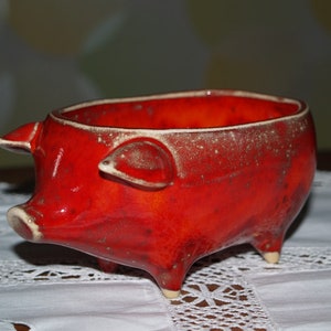May include: Red ceramic pig-shaped bowl with a speckled glaze. The bowl is sitting on a white lace tablecloth.