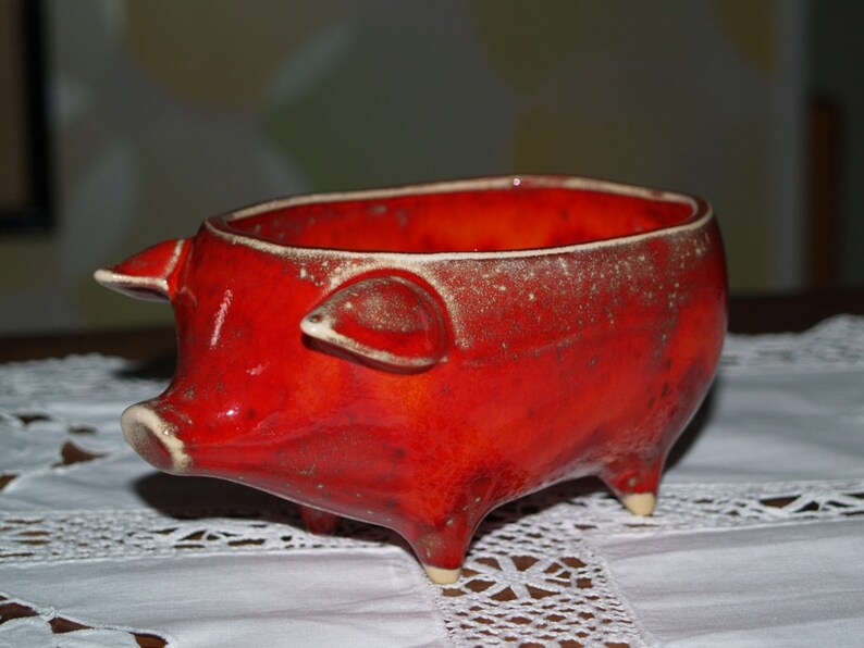 May include: Red ceramic pig-shaped bowl with a speckled glaze. The bowl is sitting on a white lace tablecloth.
