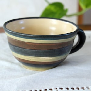 Könnte beinhalten: Eine Keramik-Kaffeetasse mit schwarzem Henkel und cremefarbenem Innenraum. Die Außenseite weist horizontale Streifen in Braun-, Blau- und Beigetönen auf. Die Tasse steht auf einer weißen Oberfläche, mit einer grünen Pflanze im Hintergrund.
