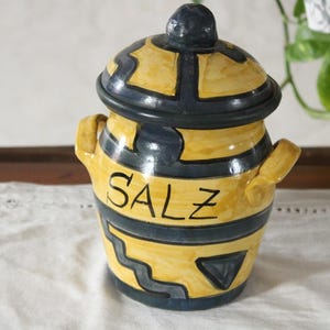 Może przedstawiać: Ceramiczna solniczka z pokrywką, pomalowana na żółto i granatowo. Słowo "SALZ" jest napisane czarnym kolorem z przodu. Słoik ma dwa małe uchwyty i dekoracyjny geometryczny wzór.