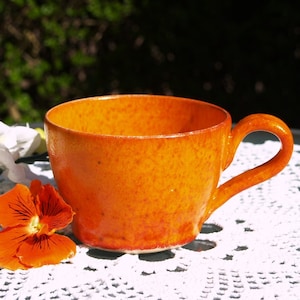 May include: A bright orange ceramic teacup with a speckled glaze sits on a white crocheted doily.