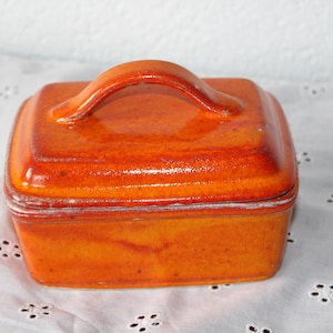 Könnte beinhalten: Orangefarbene Keramik-Butterdose mit Deckel und Griff. Die quadratische Schale hat eine glänzende Oberfläche und einen Griff zum einfachen Anheben. Die Schale hat eine warme orange Farbe und scheint handgefertigt zu sein.