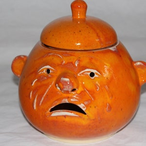 Pode incluir: Jarro de cerâmica laranja com uma tampa em forma de rosto com uma expressão triste. O rosto tem um nariz grande, olhos pequenos e uma boca larga.