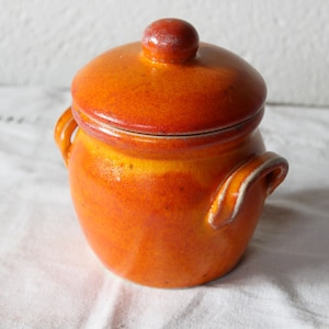 Op de afbeelding: Oranje keramische pot met een deksel en twee handvatten. De pot heeft een geglazuurde afwerking en een licht getextureerd oppervlak.