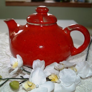Peut inclure: Une théière en céramique rouge avec un couvercle, posée sur une nappe en dentelle blanche avec des fleurs d'orchidées blanches.