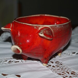 May include: A red ceramic pig-shaped bowl with white accents. The bowl is sitting on a white lace tablecloth.