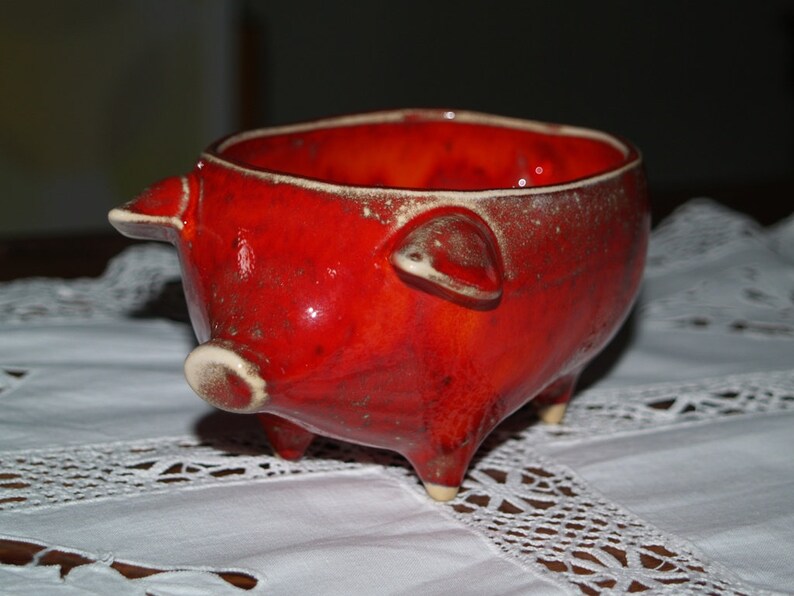 May include: A red ceramic pig-shaped bowl with white accents. The bowl is sitting on a white lace tablecloth.
