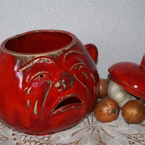 Könnte beinhalten: Ein roter Keramik-Zwiebeltopf mit einem weinenden Gesicht, mit Deckel und zwei kleinen Griffen. Der Topf steht neben ein paar Zwiebeln und einer Knoblauchzehe, alles auf einer weißen Spitzentischdecke.