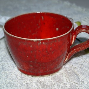 May include: A handmade ceramic mug with a vibrant red glaze. The mug features a rounded body, a sturdy handle, and a speckled texture. The rim has a contrasting off-white color, adding a touch of elegance to this kitchenware item.