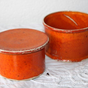 Könnte beinhalten: Zwei orangefarbene Keramikbehälter mit glänzendem Finish. Einer hat einen Deckel, der andere ist offen. Beide haben einen weißen Rand und stehen auf einer weißen, strukturierten Oberfläche. Die Behälter sind zylindrisch und scheinen handgefertigt zu sein.