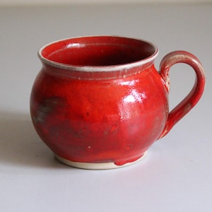 May include: A handmade, red ceramic mug with a rounded body and a curved handle. The mug has a glossy finish and a white rim. The red glaze has variations in color, adding to its unique, rustic appearance. This mug is perfect for enjoying coffee or tea.
