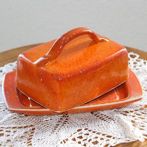 Könnte beinhalten: Orangenes Buttergefäß aus Keramik mit rechteckigem Deckel und Griff, auf passendem Unterteil. Das Gefäß steht auf einer weißen Spitzendecke und einem Holztisch. Ein Vintage-Küchenartikel.